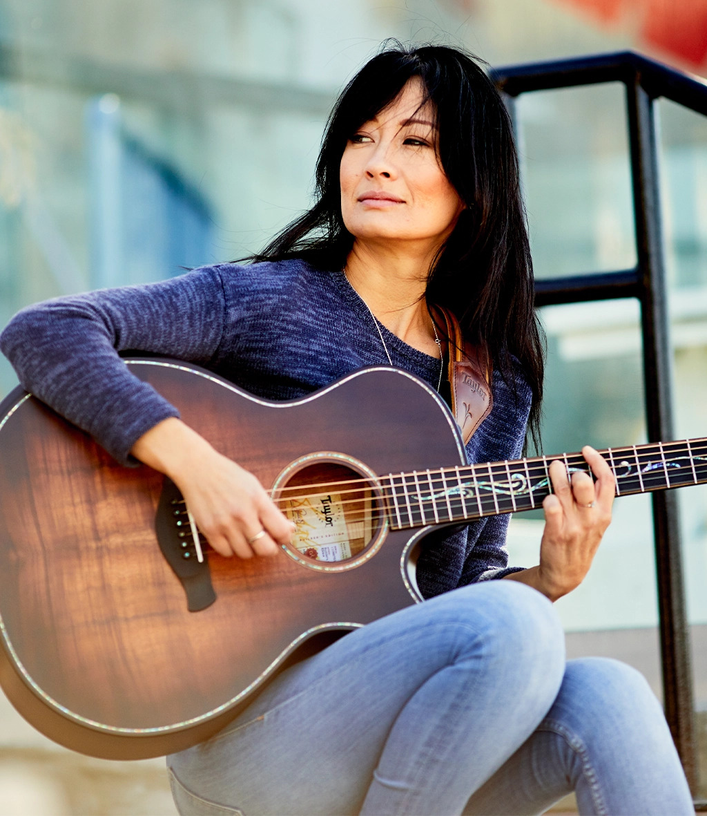 Woman staring off into distance wearing casual clothes sits down while playing guitar